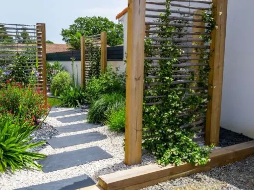 Jardin moderne avec un chemin de dalles grises. Une plante brise-vue qui pousse vite en bac grimpe sur un treillage en bois, créant un écran végétal.