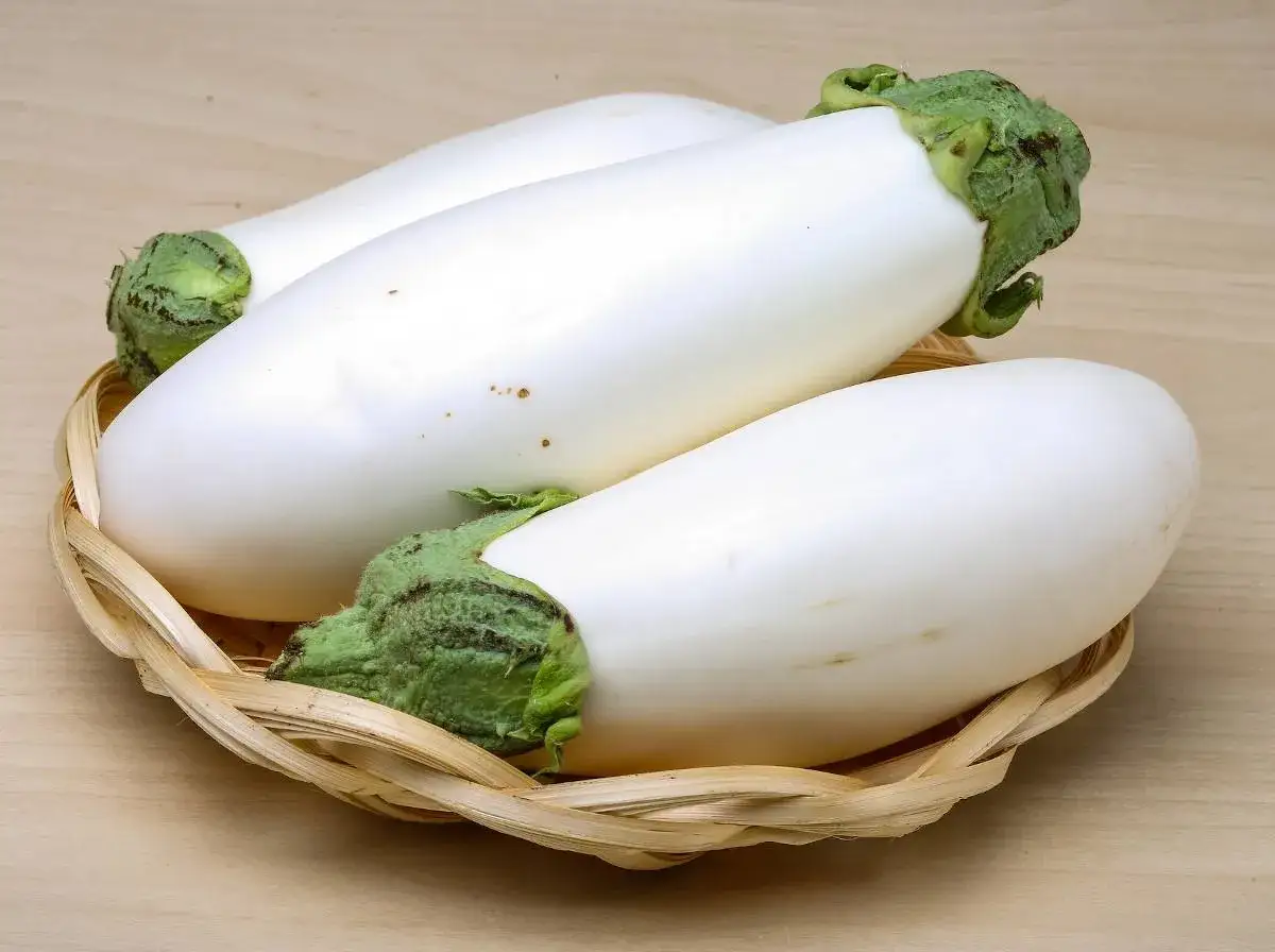 Trois aubergines blanches, d'une blancheur immaculée, reposent dans un panier en osier tressé sur une surface en bois clair.